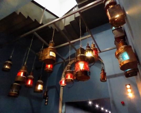 A lovely display of ships' navigation lights at the National Maritime Museum at Greenwich. Approximately 15 brass ships' lights hang from a frame near the ceiling. They display red, green, and white lights. White lights are mounted in the wall and along a corridor in the background.