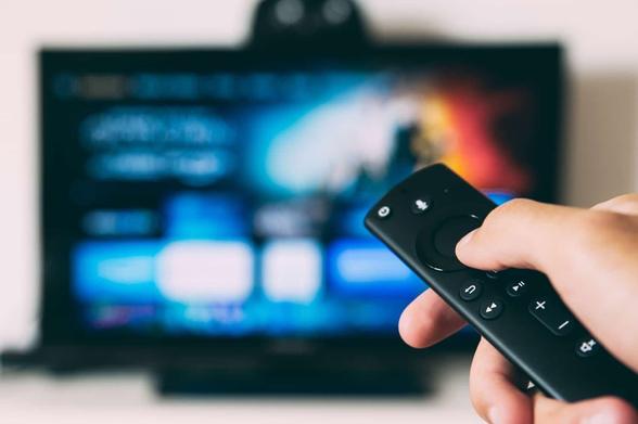 Eine Hand hält eine Fernbedienung eines Streaming Sticks in die Kamera, im Hintergrund unscharf ein Fernseher mit einer Streaming-Menüansicht.