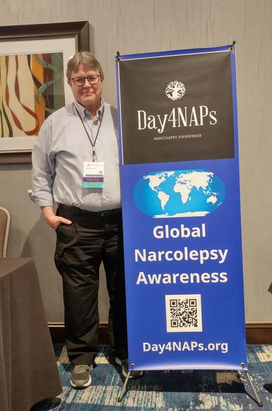 Man standing beside a banner for the narcolepsy awareness non-profit Day4NAPs.