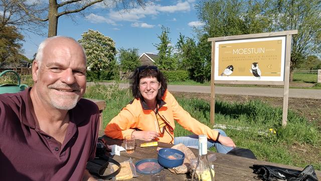 Picknick am Wegesrand, 2 Personen schauen in die Kamera, der Tisch ist mit verschiedenen Köstlichkeiten gedeckt, im Hintergrund die Werbetafel des Verweilortes