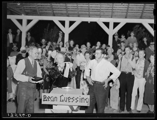 The black and white photograph depicts a group of people gathered at what appears to be an outdoor event. In the foreground, there are three individuals engaging in what seems like a game or contest known as "Bean Guessing." One person is holding up a cup containing beans, possibly for identification by taste or look, while another individual holds a book with written instructions. The third participant stands nearby observing.

The setting features a structure with wooden beams overhead, suggesting an outdoor pavilion or covered area at night time. A crowd of people surrounds the participants in various poses and expressions, indicating interest in the ongoing activity. Clothing styles suggest this event took place during mid-20th century America. The photo is labeled "Bean Guessing," which provides a clue to the nature of the game being played.

The image's context indicates it was taken at Shafter migrant camp located in California, possibly around Halloween time given the label on the photograph and the festive atmosphere among attendees.