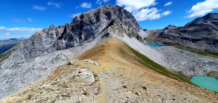 Foto einer Berglandschaft. Im Vordergrund ein Grat aus gelb-orangem Gestein, dahinter ein Gipfel und rechts zwei Bergseen.