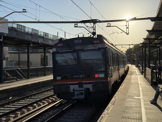 Se ve la cola de un automotor eléctrico de la serie 470 pasando por la estación de Sitges, en vía doble, ligeramente tumbado por estar en una zona en curva.

El poste de la catenaria compartida entre varias vías tapa parcialmente el sol, en el atardecer.