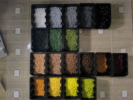 A view of the 30 clear pegboards, 5 by 6, arranged on my desk. On top of the boards there are 18 boxes with different colored beads. There are black, white and grey, green, brown and yellow beads.