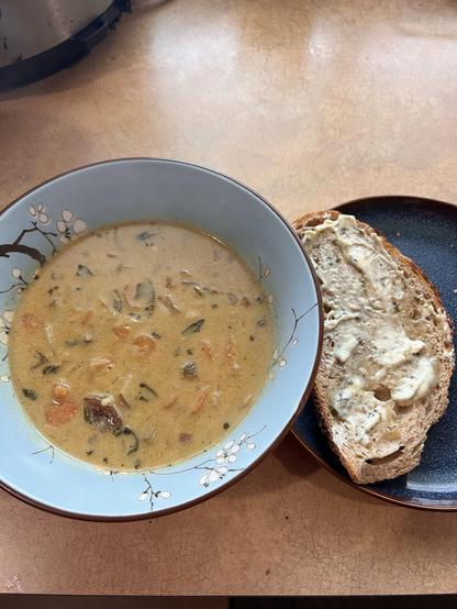 Photo of a blue bowl full of an orangish stew with bits of herbs, carrots, and potatoes visible. A slice of bread with an herb butter melting on top is posed on a plate to the side of the bowl.