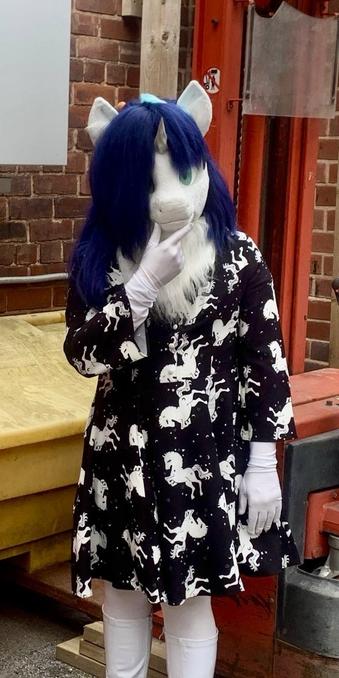 Light (unicorn fursuit, white fur with blue mane) stands in front of a forklift in the garden of a pub. She is wearing a black dress with a white unicorn pattern and in a thoughtful pose.