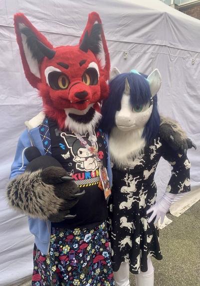 Light (unicorn fursuit, white fur with blue mane) and Lillith (devil fox fursuit) stand in a hug in front of a large white marquee.