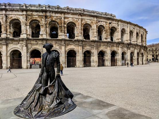 A colour photograph shows a bronze statue of a bullfighter in the foreground, set against the backdrop of the ancient Roman amphitheatre in Nîmes, France. The statue depicts a matador in traditional attire, holding a large cape that flows around him. The amphitheatre is a large, oval structure made of weathered stone, featuring two tiers of arched openings. Several people can be seen as small figures in the open plaza in front of the arena, giving a sense of scale to the massive building. The sky above is partly cloudy.