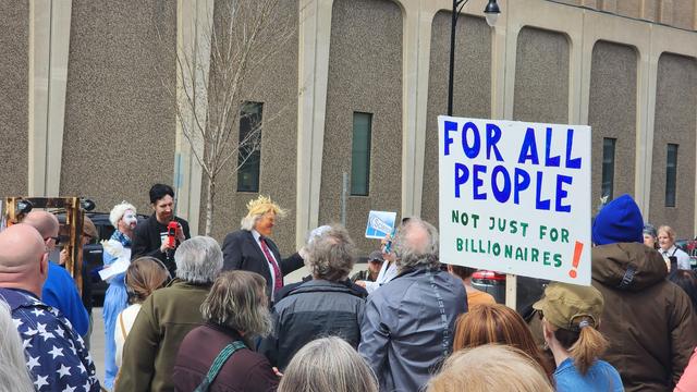 A crowd of people, a sign saya For All People Not Just For Millionaires. A Trump actor lookalike wears a gold crown.