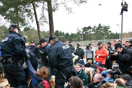 Viele Menschen auf dem Boden vor einem stabilen Metallzaun. Im Vordergrund etliche Polizist:innen in blauer Uniform. Im Hintergrund ein Hubschrauber. Eine Person klettert an einem Laternenmast.