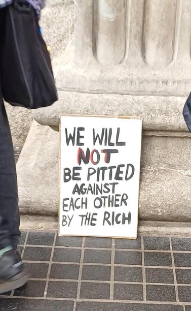 Cardboard handmade sign rests against a pillar of the GPO, reading "We will not be pitted against each other by the rich"