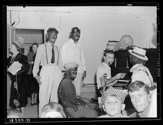 The image is a black and white photograph depicting an indoor gathering with multiple individuals. Some people are dressed in costumes that mimic characters or personas, possibly for entertainment purposes such as Halloween festivities. The subjects include men wearing hats, overalls, suspenders, and makeshift masks to resemble different figures; these outfits suggest they have engaged in creative costume-making activities.

The setting appears informal, likely a communal space with various items like books on shelves visible in the background. A child is also present among the adults, observing or participating in the event. The atmosphere seems lively yet cozy as if capturing candid moments during an impromptu gathering for amusement and socialization.

Additional context provided indicates that this could be related to Camp Talent providing music at Shafter camp for migrants on Halloween. This setting further implies a historical moment where such celebrations were part of community-building activities amidst migration challenges in California, specifically highlighting the inclusivity within these camps through cultural events like talent shows or costume parties.