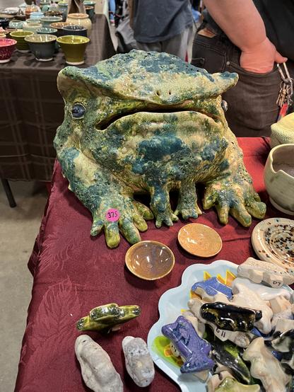 A basketball-sized ceramic frog. It’s got splotchy green and yellow colors with a properly bumpy shiny skin. It’s back legs are way bigger than the two little front legs and its mouth is wide (and hollow). It has a $375 price tag, and is sitting on a display table along with other (much smaller) ceramic items
