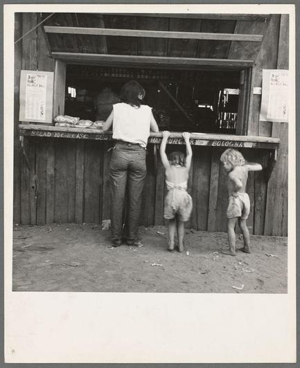 The image is a black and white photograph depicting an outdoor scene. In the center, there's a woman standing in front of a makeshift food stand with shelves holding various items like "BREAD," "ICE-CREAM SODA," and other products labeled such as "CHARGE $2 FOR BOLONA." The text on these signs is not entirely clear.

The woman appears to be wearing casual work attire, including a short-sleeve shirt and pants. Her hair seems pulled back, suggesting practicality for her job or activity. She has both hands gripping the top of two children who are reaching up towards the food stand's counter. The older child on the left is standing with their legs slightly apart while lifting themselves to look at what might be inside a display case within the stall.

The younger child on the right appears smaller in stature, wearing shorts and looking upward as if trying to get an even better view of whatever they are interested in from behind the counter. Both children have bare feet, which indicates a setting where shoes were not required or it is too warm for footwear.

Behind them stands a wooden fence that suggests this might be at some sort of outdoor market or event rather than within a typical urban environment. The ground appears to be dirt with small debris scattered around, and the general atmosphere feels rustic and down-to-earth.

In terms of context provided in the source link description, it seems related to hop picking work  [...]