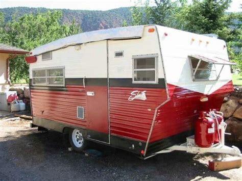 A vintage-style travel trailer in red and white, featuring multiple windows and a distinctive shape. It is parked outdoors, with greenery and mountains in the background.