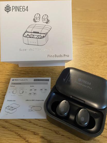 Wireless bluetooth earbuds sitting in a black charging case. The case lid is slid open revealing the earbuds inside. Case and earbuds are a dark grey/charcoal color. The case lid has Pine64 printing on it. The earbuds have a small pine64 logo (pine cone). Instruction booklet is in frame to the left of the case. PineBuds retail box is in view above the case. The items are placed on a light wood counter top that has dark grain.
