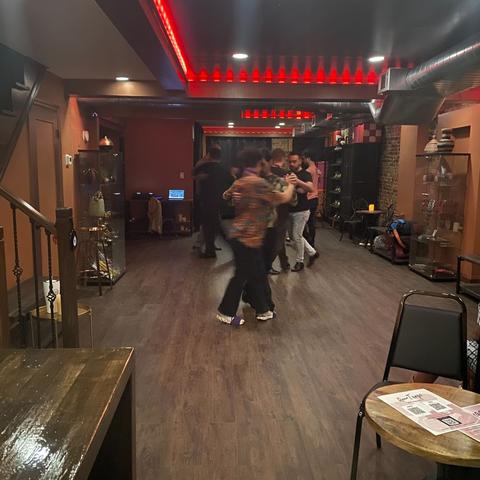4 couples whirl around a wooden dance floor lined with display cases of Argentine mates and artwork. There is red lighting along the ceiling beams.
