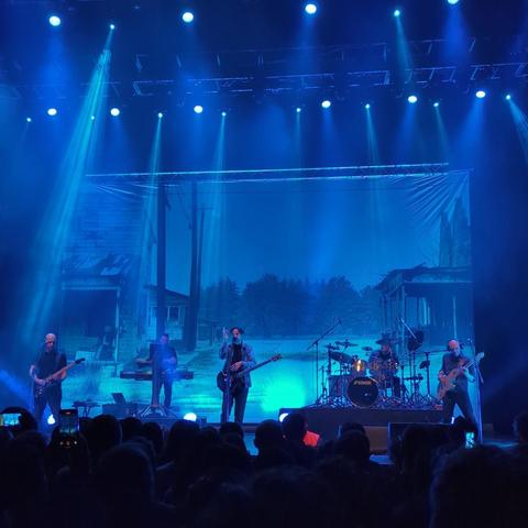 La banda The Pineapple Thief toca en un escenario iluminado en azul. Detrás de ellos hay una gran pantalla que muestra una imagen de un paisaje rural con casas viejas y árboles. La banda consta de cuatro miembros: un guitarrista principal, un bajista, un baterista y un tecladista. El público está sentado frente al escenario, parcialmente visible. La atmósfera es oscura, excepto por la iluminación azul intensa centrada en la banda.