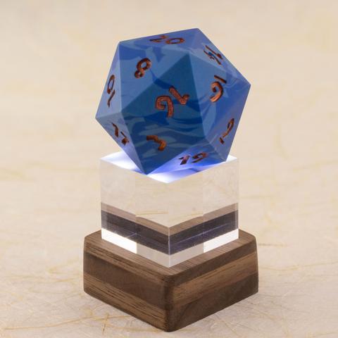 A one-inch clear acrylic cube sits in a small square layered walnut stand, which lights the cube and the 42 millimeter 20-sided die sitting on top of it from below. The die is a swirl of two shades of blue with metallic copper numbers.  Shown against a defocused background of handmade paper with fiber inclusions. Side view.