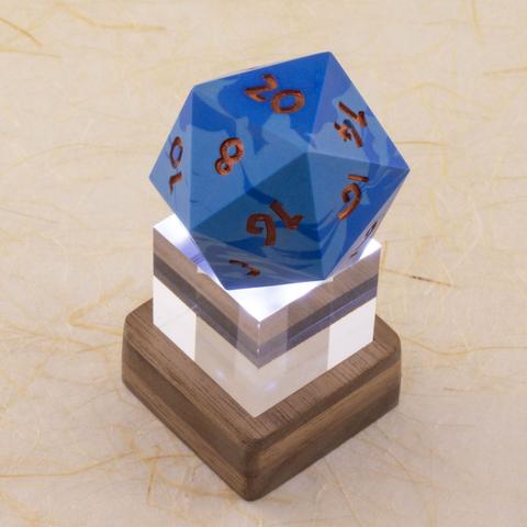 A one-inch clear acrylic cube sits in a small square layered walnut stand, which lights the cube and the 42 millimeter 20-sided die sitting on top of it from below. The die is a swirl of two shades of blue with metallic copper numbers.  Shown against a defocused background of handmade paper with fiber inclusions. Three-quarter top-down view.