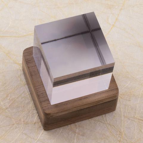 Three-quarter top-down view of a one-inch clear acrylic cube sitting in a small square layered walnut stand. The stand lights the cube from below, but the light is off in this image. Shown against a background of handmade paper with fiber inclusions.