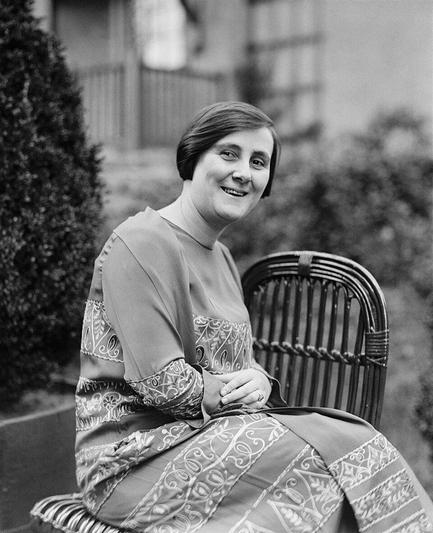 Portrait en noir et blanc de Bertha Lutz pris le 27 avril 1925. Elle est assise sur une chaise en osier, portant une robe décorée de motifs complexes et élégants. En arrière-plan, on distingue un jardin avec des buissons et une structure architecturale.