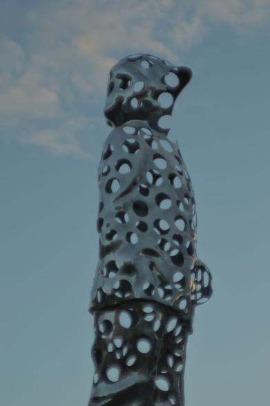 A abstract statue of a human standing up, covered in holes that let the evening blue sky pass through.