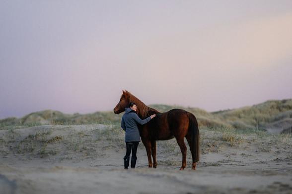 A horse and their human in the dunes during dawn. The woman is standing next to the horse's shoulder and gently touching it's back while looking back. The horse is standing proud and aware while looking in the opposite direction.