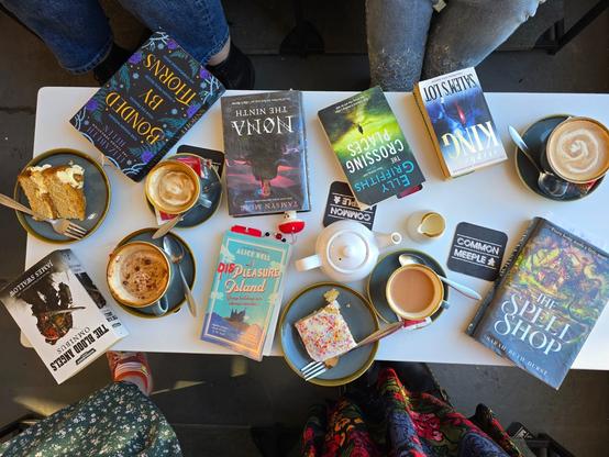 An overhead view of a low white table crammed with seven varied fiction books, three cups of coffee, a cup of tea (with teapot) and two slices of cake. We can just see four pairs of legs, two pairs wearing jeans, two pairs wearing skirts, at the edges of the table. My legs are encased in the paler pair of jeans which, coincidentally, I am also wearing today.