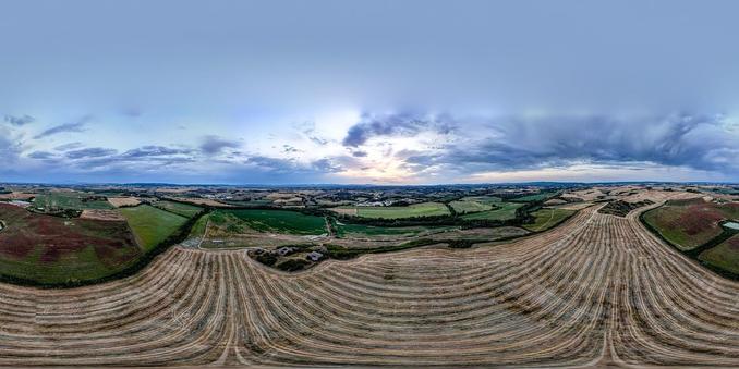 This image is a panoramic view of a vast landscape, showing extensive agricultural fields with furrows likely created during the plowing or harvesting process. The landscape includes varying patterns and shades in the fields, suggesting different types of crops or stages of cultivation. The background is adorned with a dramatic sky where clouds are scattered, suggesting either dawn or dusk due to the soft lighting. There are patches of greenery, likely patches of grass or other vegetation, which indicate some sections of the land are not used for agriculture or are in different phases of the crop cycle. The undulating terrain contributes to the depth and richness of the scene.