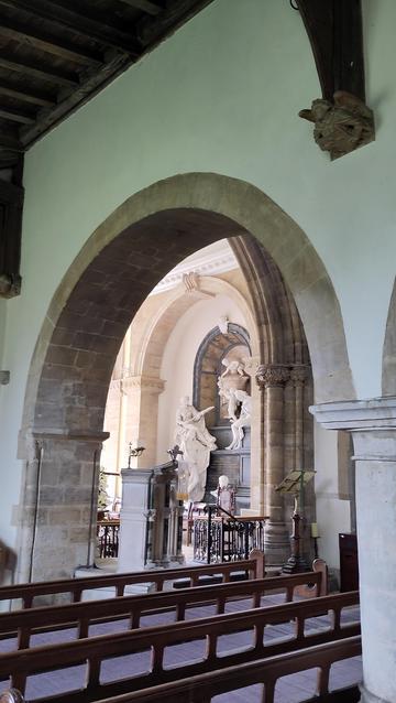 Looking through plain rounded archway, can see the edge of a more decorative pillar and arch, and beyond that a Georgian style interior with alabaster statuary.