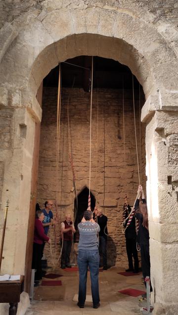 A chunky rounded archway on top of tall square section walls, framing a group of rongers using ropes with woollen sallies to ring the bells.