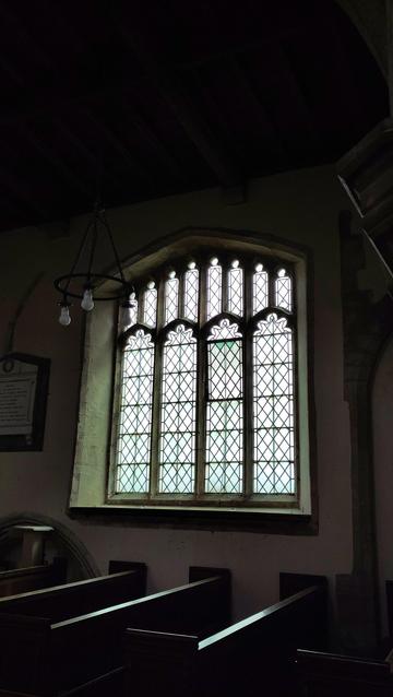 Looking from the inside at a perpendicular style church window, so the framework is silhouetted against the light. The top arch is very angular. It has 4 main sections which split into 8 sections at the top.