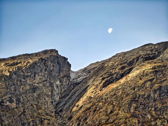 This image portrays a rugged mountain landscape under a clear sky with the moon visible in the background. The mountains have steep and rocky slopes with a variety of earthy tones from dark browns to golden hues, likely highlighted by the low angle of sunlight. The visible geological features and erosion patterns suggest a harsh and possibly arid environment. The presence of the moon adds a serene, almost otherworldly quality to the scene.
