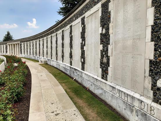 This image shows a war memorial with a long curved wall containing the names of fallen soldiers inscribed on stone panels. The phrase "COMRADES IN DEATH 1918" is visible, indicating that the memorial commemorates individuals who died in 1918, possibly during World War I. The wall is surrounded by neatly maintained grass and a flowerbed with roses. The design and maintenance of the memorial provides a solemn, respectful space to honor the lives of those listed.