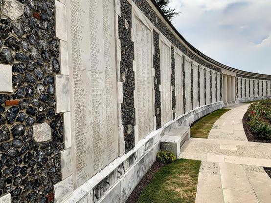 This image shows a war memorial. The structure consists of a long, curving wall featuring numerous names etched into its surface, presumably commemorating individuals who have died in a military conflict. The wall is made of panels interposed with sections of flint aggregate—a traditional and often-used material in memorial architecture that conveys solemnity and resilience. There is a walkway along the base of the wall, and a well-maintained grass lawn and manicured flower beds add a serene touch to the setting. The design of the memorial, with its open colonnade in the background, suggests a peaceful place for reflection and remembrance.