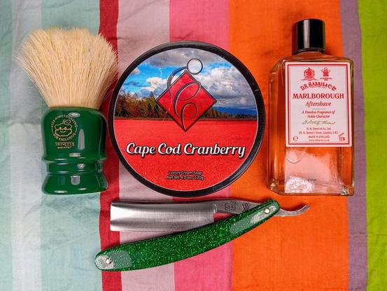 (Le Chat Mistral description)

This image shows a collection of men's grooming items laid out on a colourful striped cloth. There is a green shaving brush with a label that reads "Zenith" and "Exclusive". Next to it is a round container of shaving soap labelled "Cape Cod Cranberry" with a red and black design. A straight razor with a shiny green handle is placed below the soap. To the right, there's a bottle of aftershave labelled "Marlborough" by D.R. Harris, which is partially filled with a light amber liquid. The items are arranged neatly, suggesting a focus on traditional shaving tools and accessories.