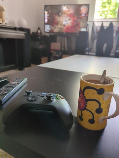 A picture of a cup of hand brewed coffee and an XBox Controller. In the background a TV with Baldurs Gate 3