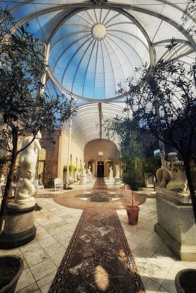 A wide-angle shot inside a bright conservatory with a glass dome roof, featuring marble floors, a central patterned walkway, various potted trees, and several white classical sculptures.