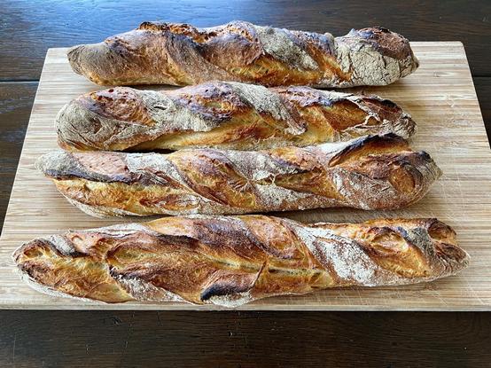 Auf einem dunklen Holztisch liegt ein Holzbrett. Darauf liegen vier frisch gebackene Baguette mit rustikalem Ausbund.