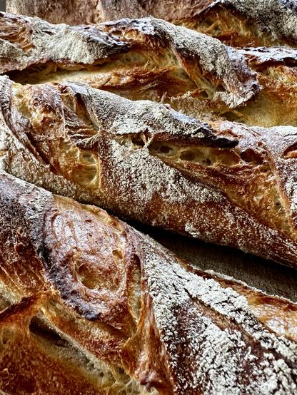 Auf einem dunklen Holztisch liegt ein Holzbrett. Darauf liegen vier frisch gebackene Baguette mit rustikalem Ausbund.