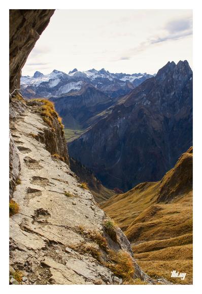 A narrow high-alpine path leading over a precarious rocky ledge with improvised, carved out footsteps and secured with steel ropes. Next to it, steep meadows with dried out golden brown grasses drop off 700-800m into the valley below. Layers of major snow capped mountains in the background. Semi blown out bright sky.