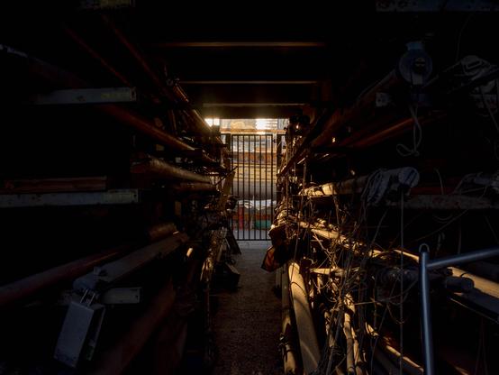 Photo looking through a dark storage area into a boat yard. Through a tall metal gate at the far end can be glimpsed a wooden boat in afternoon sunshine, the light spills in from that end, through a tall metal gate, and partially highlights a confusion of rigging and masts, all laid down lengthwise on shelves down either side.