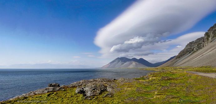 A colour photo of a seascape under a mostly clear deep cyan sky. The viewpoint is coastal grassland dotted with boulders and a stretch of still blue sea on the left. The mountains rising from scree slopes on the right have layered and rounded clouds hanging above them, casting shadows onto the bare rocky flanks. It's oddly beautiful.