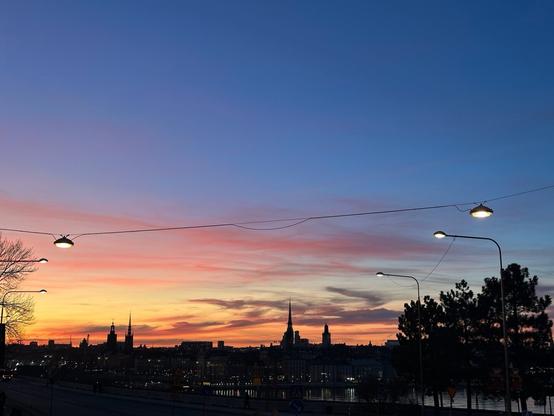 the silhouette of a city backlit by the sunset. There are wisps of cloud, and a string of street lamps hangs in the foreground