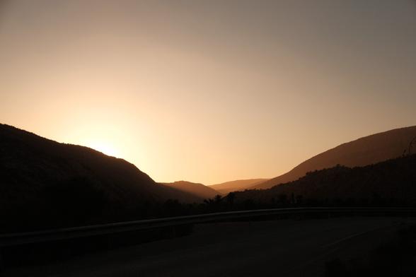 A scenic view of mountains bathed in the warm, golden light of the setting sun. A road with a guardrail runs along the foreground, shadowed by the light. Dim silhouettes of palm trees and other foliage are visible nestled among the hills. The sky has a soft, gradient tone from yellowish-oranges near the horizon up to a lighter color, creating a peaceful, atmospheric effect.