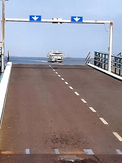Auf dem Bild sieht man den Fähranleger in Den Helder (Niederlande), von dem regelmäßig die Fähren zur Insel Texel an- und ablegen. Im Hintergrund ist das ankommende Schiff zu erkennen, das in wenigen Minuten an dem Steg anlegen wird. Über der Straße weisen zwei blau-weiße Schilder auf die zweispurige Auffahrt auf die Fähre hin.