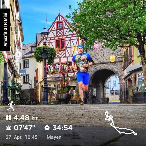 Das Bild zeigt einen Läufer, der durch eine malerische Stadt läuft. Der Läufer trägt ein blau-grünes Outfit und ist in der Bewegung festgehalten. Im Hintergrund sieht man ein traditionelles deutsches Fachwerkhaus mit Steinbogen und eine Baumkrone auf der rechten Seite. Links befindet sich eine Straßenlaterne. Außerdem sind Laufdetails eingeblendet: ‚Amazfit GTR Mini‘, eine Icon-Grafik mit ‚4,48 km‘, ‚07'47"‘, ‚34:54 Laufzeit‘ und das Datum ‚27. Apr., 10:45 | Mayen‘. Unten rechts zeigt eine kleine Karte die Laufroute.