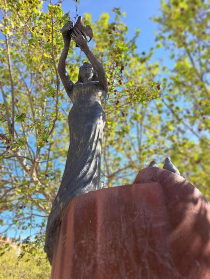 Detalle de la escultura realizada en bronce, una mujer sostiene a una paloma con los brazos estirados hacia el frente y un poco por encima de la altura de su cabeza. 

La escultura muestra mujeres a tres alturas y esta es la que se encuentra en lo más alto.

Bajo ella está el pedestal que sustenta la obra en sus varios niveles, es de acero corten.

A la derecha hay dos palomas sobre el pedestal.