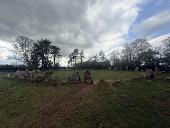 A circular arrangement of stone monoliths is set against a backdrop of green grass and a cloudy sky, with trees surrounding the area.